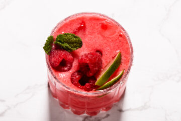 A glass of bright pink raspberry drink with ice, garnished with fresh raspberries, lime wedges, and mint leaves on a white surface.