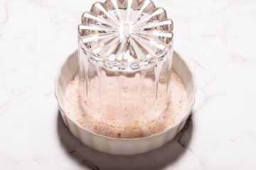 A clear glass is pressed upside down into a round dish filled with pink salt on a white marble surface.