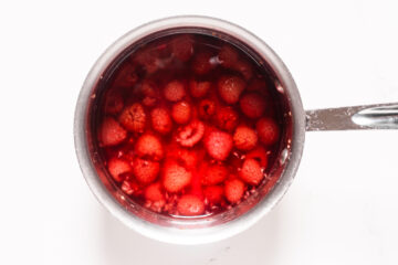 A saucepan filled with raspberries submerged in red liquid, viewed from above on a white surface.