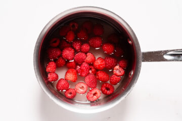 A stainless steel saucepan filled with water and fresh raspberries, viewed from above on a white surface.