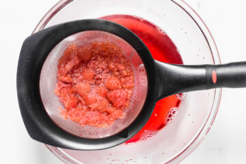 A fine mesh strainer holding mashed strawberries over a glass bowl, with red juice collected in the bowl below.