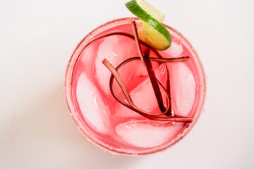 Top-down view of a pink drink with ice cubes, garnished with rhubarb strips and a lime wedge on the rim of the glass.
