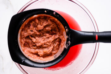 A black strainer holds mashed fruit pulp over a glass bowl containing red juice, viewed from above.