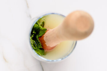 Top view of a wooden muddler pressing mint leaves in a glass containing a light-colored liquid, on a white surface.