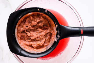 A fine mesh strainer holding fruit pulp sits over a clear bowl containing red juice.