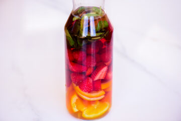 A glass bottle filled with dark liquid, containing orange slices, strawberries, raspberries, and fresh mint leaves.