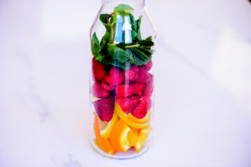 A glass bottle filled with slices of orange, strawberries, raspberries, and fresh mint leaves, placed on a white surface.