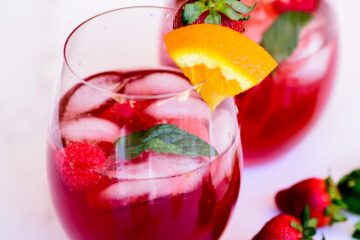A glass of red iced drink garnished with a mint leaf, orange slice, raspberry, and strawberry, with ice cubes and additional strawberries nearby.