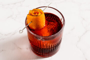 A glass of red cocktail with a large ice cube, garnished with an orange peel twist on a metal pick, set on a white marble surface.