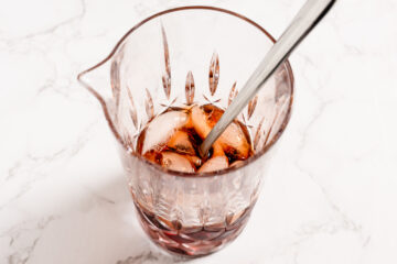 A glass mixing pitcher with a metal stirring spoon, ice cubes, and a dark liquid, placed on a white marble surface.