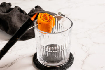 A glass with a decorative orange peel garnish on a skewer, being flamed with a lighter, sits on a dark coaster against a white marble background.
