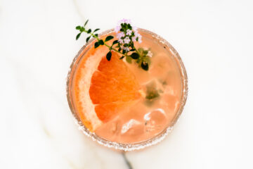 A glass with a salted rim, filled with ice, a pink beverage, a grapefruit slice, and garnished with a small sprig of flowers and herbs.