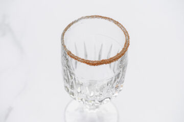 A clear, empty glass with a faceted design and a rim coated in brown sugar crystals sits against a white background.