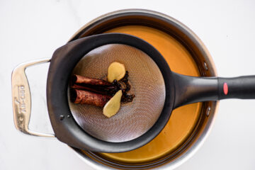 A mesh strainer with cinnamon sticks, star anise, and ginger slices sits over a pot of amber-colored liquid.