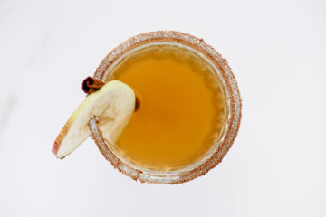 Overhead view of a glass with a brown beverage, garnished with a slice of apple and a cinnamon stick, and a rim coated with sugar and spices.