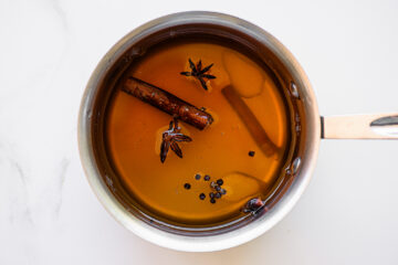 A saucepan filled with amber-colored liquid, containing a cinnamon stick, star anise, and whole black peppercorns, sits on a white surface.