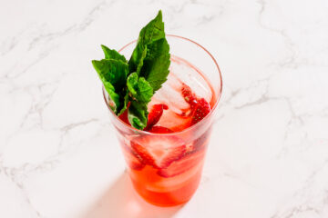 A glass of iced strawberry drink garnished with fresh mint leaves, placed on a white marble surface.