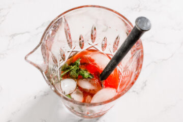 A glass mixing pitcher with ice, mint leaves, and a red liquid, stirred with a black stirring stick, on a white marble surface.