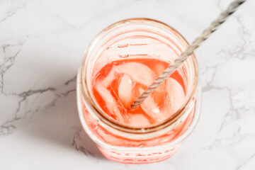 A glass jar filled with a pink beverage and ice cubes, with a metal stirrer inside, on a white marble surface.