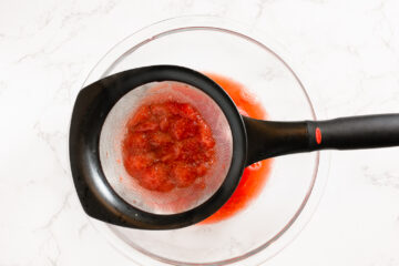 A fine mesh strainer with crushed strawberries sits over a glass bowl, separating juice from pulp on a white countertop.