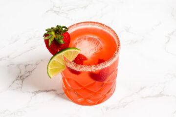 A red drink in a glass with a sugared rim, garnished with a strawberry and lime slice, served over ice on a white marble surface.