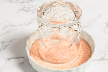 A glass cup is placed upside down in a shallow white dish filled with pink rimming salt on a marble surface.