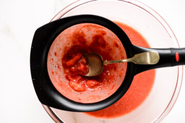 A metal spoon presses strawberry pulp through a mesh strainer into a glass bowl filled with strawberry juice.