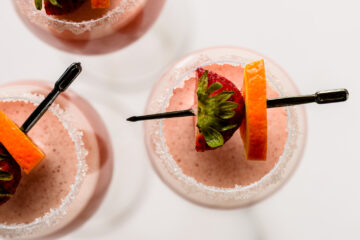 Three glasses of pink blended drinks with sugared rims, each garnished with a strawberry and an orange slice skewered on a black cocktail pick, viewed from above.