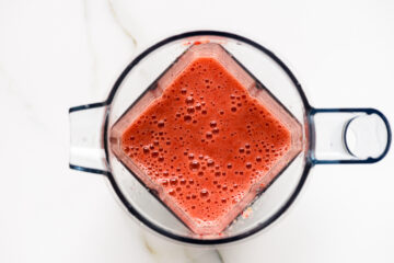 A top-down view of a blender filled with a frothy red smoothie on a white surface.