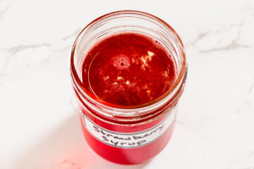 A jar filled with red strawberry syrup, labeled strawberry syrup, on a white marble surface.
