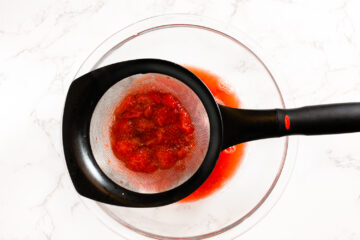 A fine mesh strainer with crushed strawberries sits over a glass bowl, collecting juice while pulp remains in the strainer.