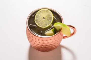 A copper mug filled with a dark beverage, ice, and garnished with a lime wheel and lime wedge on the rim, set on a white surface.