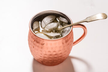 A copper mug filled with ice cubes and a drink, with a metal spoon inside, set on a white surface.