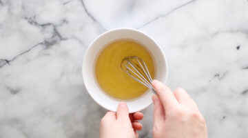 A person uses a whisk to mix a yellow liquid in a white bowl on a marble countertop.
