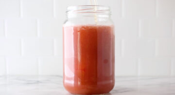 A large glass jar filled with pink juice sits on a marble surface with a straw inside, against a white tiled background.