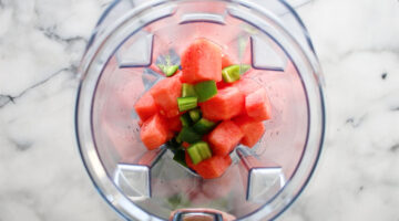 Chunks of watermelon and pieces of green bell pepper in a blender, viewed from above on a marble countertop.