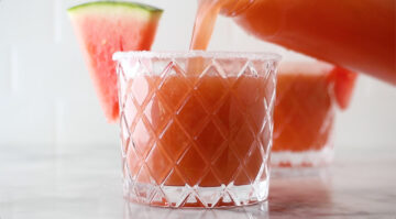 A crystal glass with a salted rim is being filled with a pink watermelon drink, garnished with a slice of watermelon.