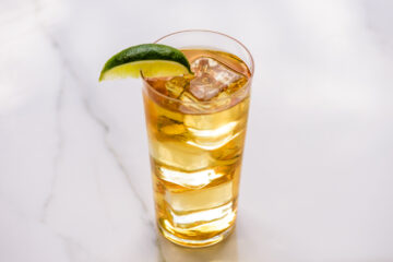A tall glass filled with a golden-brown beverage, ice cubes, and garnished with a lime wedge, placed on a white marble surface.