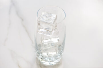A clear glass containing three ice cubes, placed on a white marble surface.