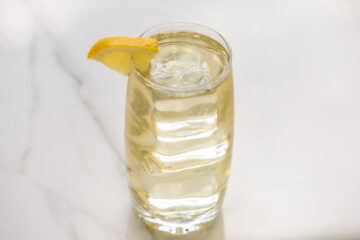 A tall glass of a light-colored beverage with ice cubes and a lemon wedge garnish on a white surface.