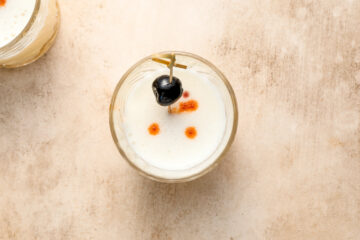 A glass of frothy cocktail garnished with a cherry and a toothpick, topped with several drops of bitters, on a light-colored surface.