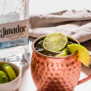 A copper mug filled with a cocktail garnished with lime slices sits next to a bottle of El Jimador tequila and a small bowl of lime wedges on a white surface.