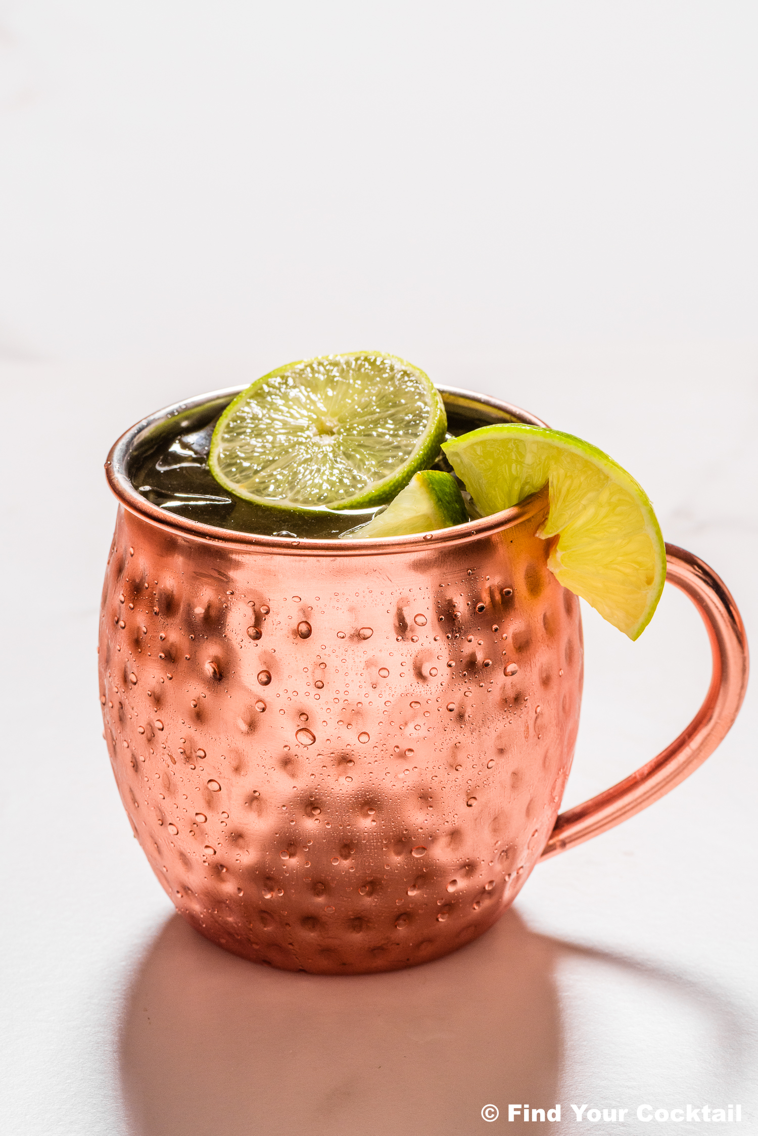A copper mug filled with a drink, garnished with two lime wedges, on a white surface.