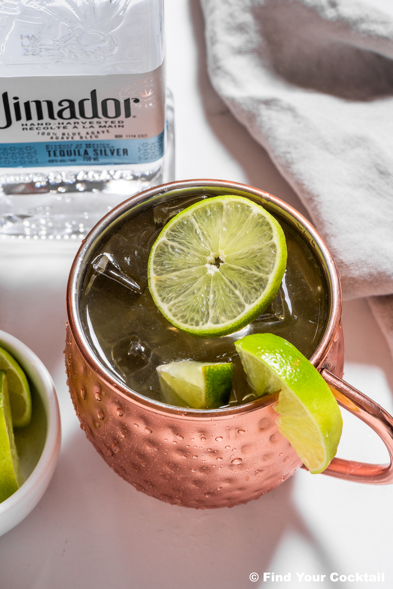 A copper mug filled with a cocktail, ice, and lime slices, sits on a white surface next to a bottle of El Jimador tequila and a bowl of lime wedges.