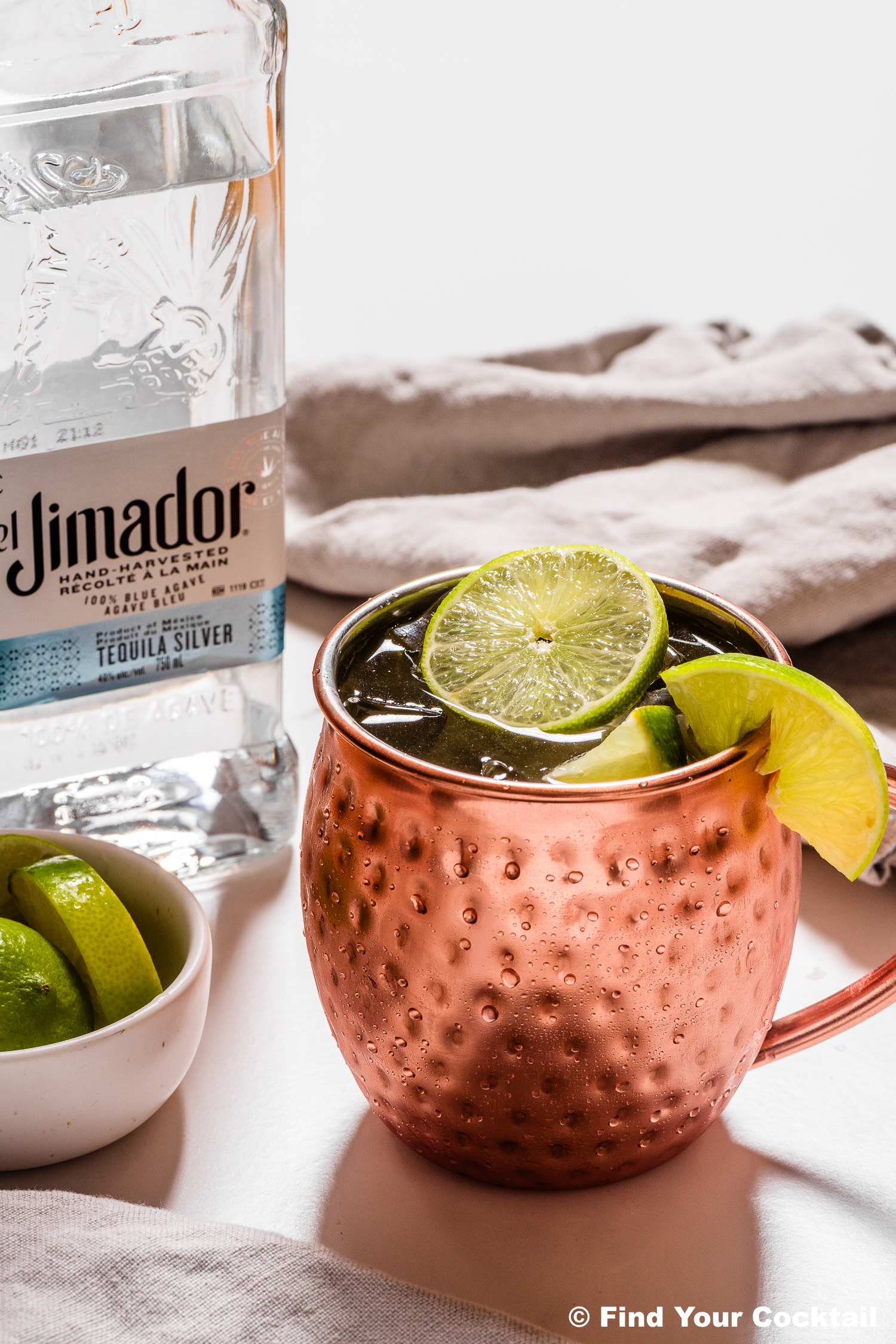 A copper mug filled with a cocktail garnished with lime slices sits next to a bottle of El Jimador tequila and a small bowl of lime wedges on a white surface.