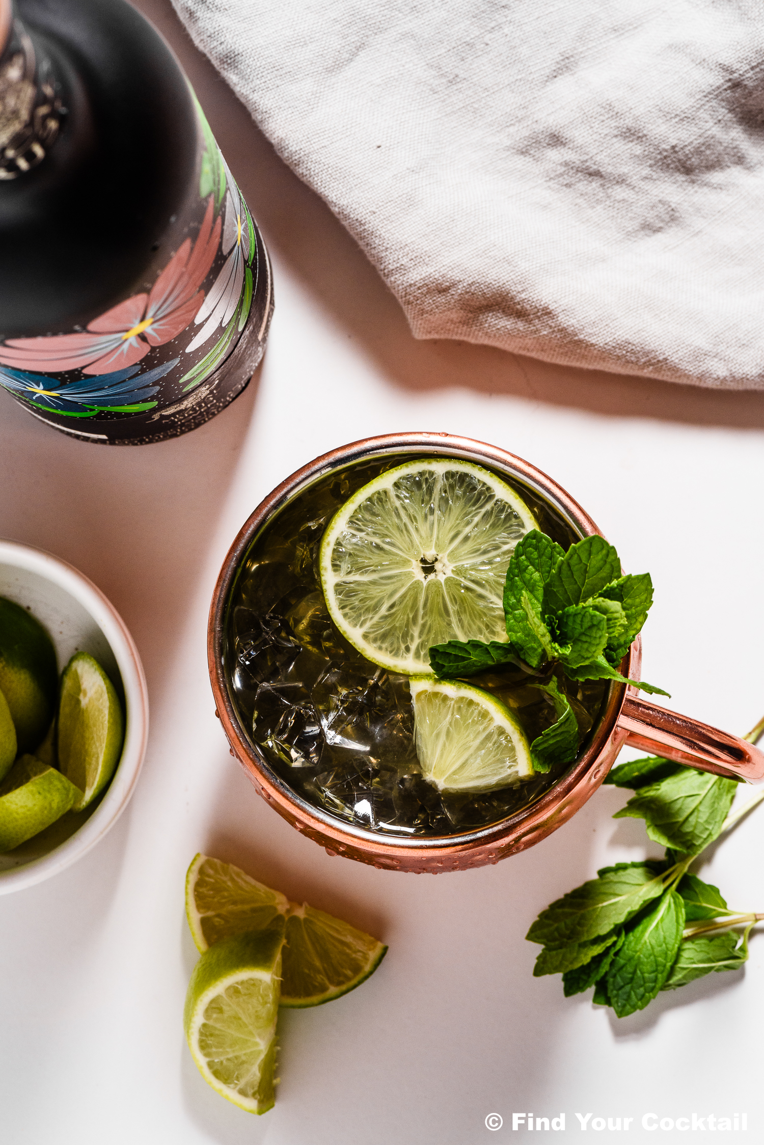 A copper mug filled with a cocktail garnished with lime slices and mint, surrounded by lime wedges, a white bowl of limes, and a floral-patterned bottle on a white surface.
