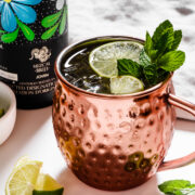 A copper mug filled with a cocktail garnished with lime slices and mint, next to a bottle with a floral design and lime wedges on a white surface.