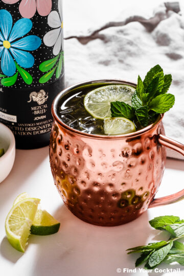 A copper mug filled with a cocktail garnished with lime slices and mint, next to a bottle with a floral design and lime wedges on a white surface.