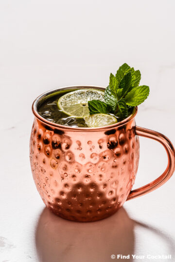 A copper mug filled with a dark beverage, garnished with two lime slices and a sprig of mint, placed on a light surface.