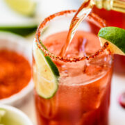 A glass rimmed with chili powder, filled with ice and a reddish drink, is garnished with lime slices while another beverage is being poured in.